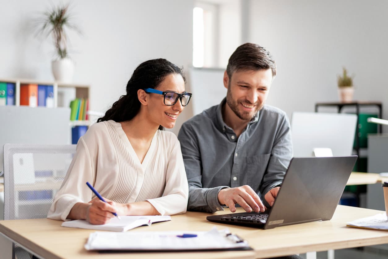 Two colleagues working together in a bright office environment, sharing a laptop