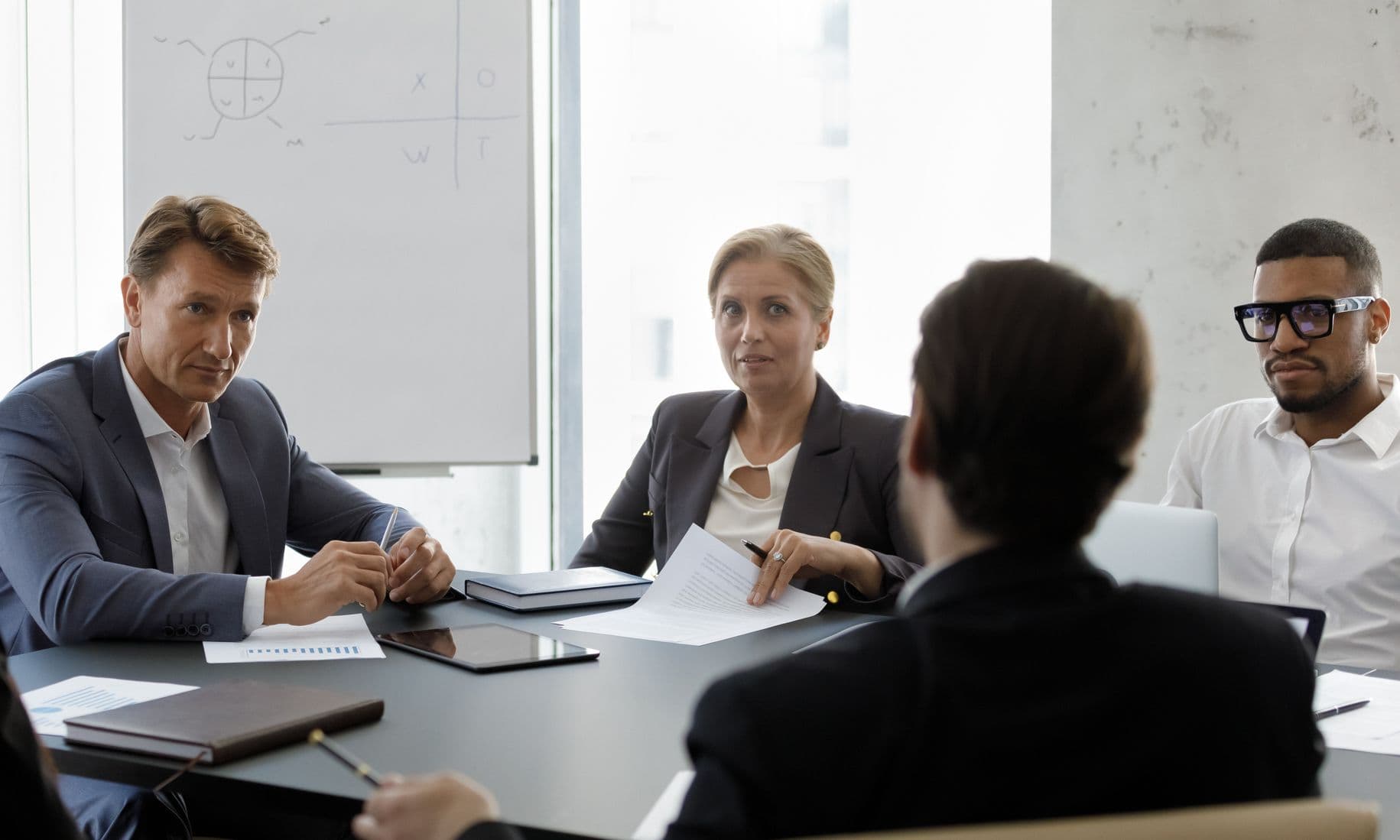 Four executives sit around a boardroom table during a meeting, with one person’s back to the camera